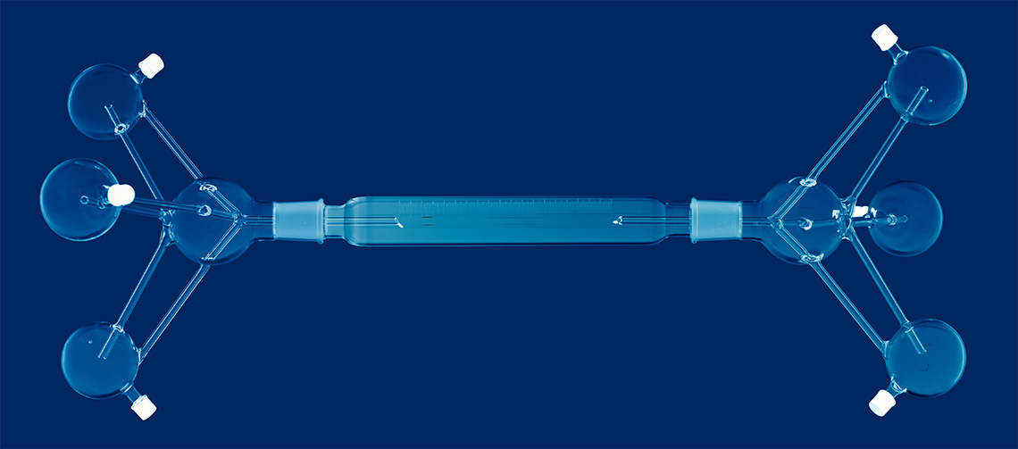 The Reversible Drip Device (RDD), here shown horizontally rather than upright, 2024. Based on an hourglass, the RDD is used to measure the viscosity and drip-length of saliva and artificial saliva types used in Utter. The long, graduated cylinder in the center can measure spit driblets up to 30 cm. It was completed by Paul Vanouse and Tracy Drier during a weeklong residency at Salem Community College Glass Education Center.
